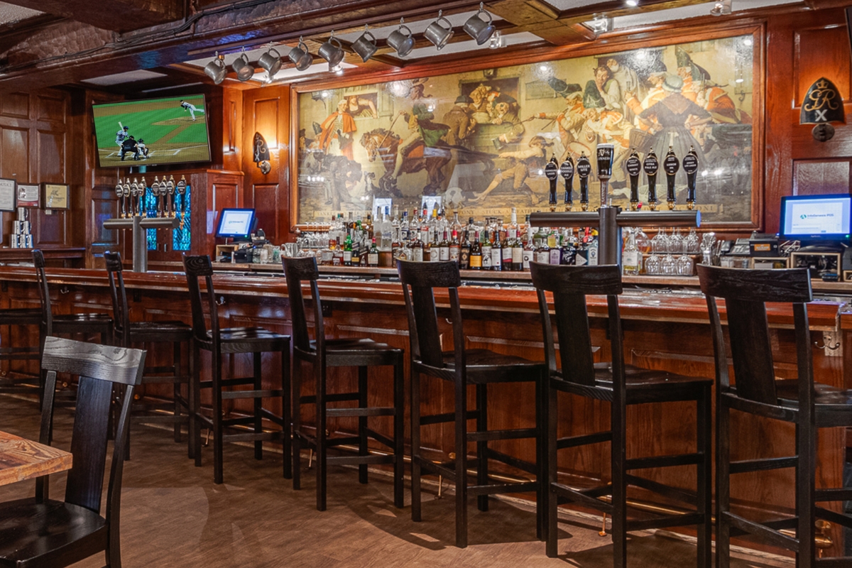 Image of the bar at The Nassau Inn (1756), a member of Historic Hotels of America since 2024. Pictured a long wood bar with stools in front of an original mural.
