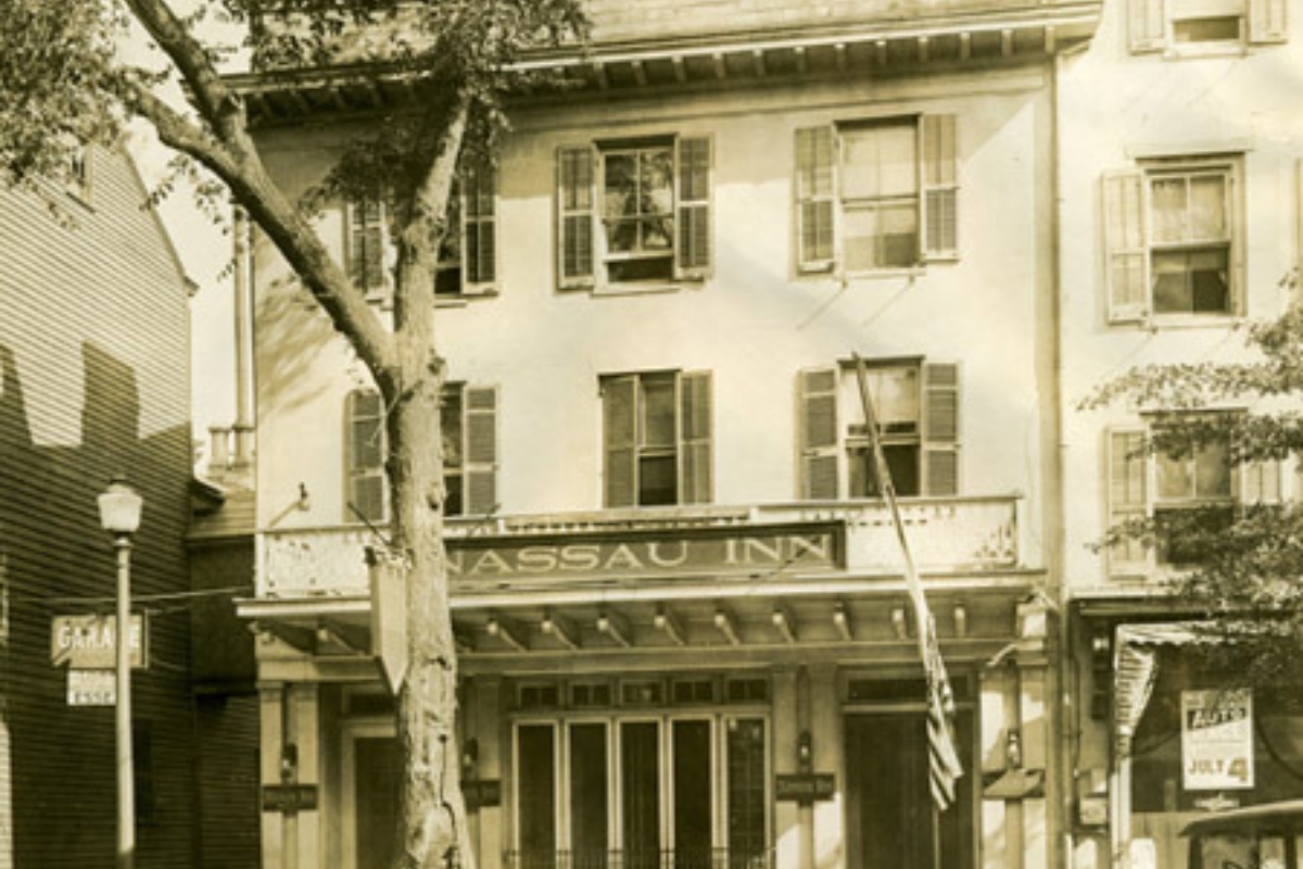 Historic image of the original exterior of the Nassau Inn (1756), a member of Historic Hotels of America since 2024. Pictured is the front of the building with several flags and signs.