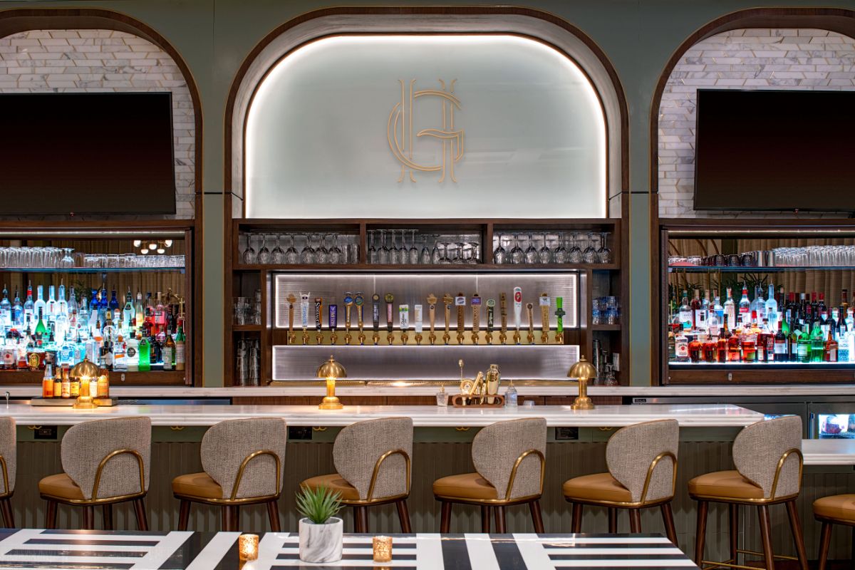 Image of a bar at Hilton Richmond Downtown (1885), a member of Historic Hotels of America since 2024. Pictured is a bar with stools and bottles and glasses beyond it.