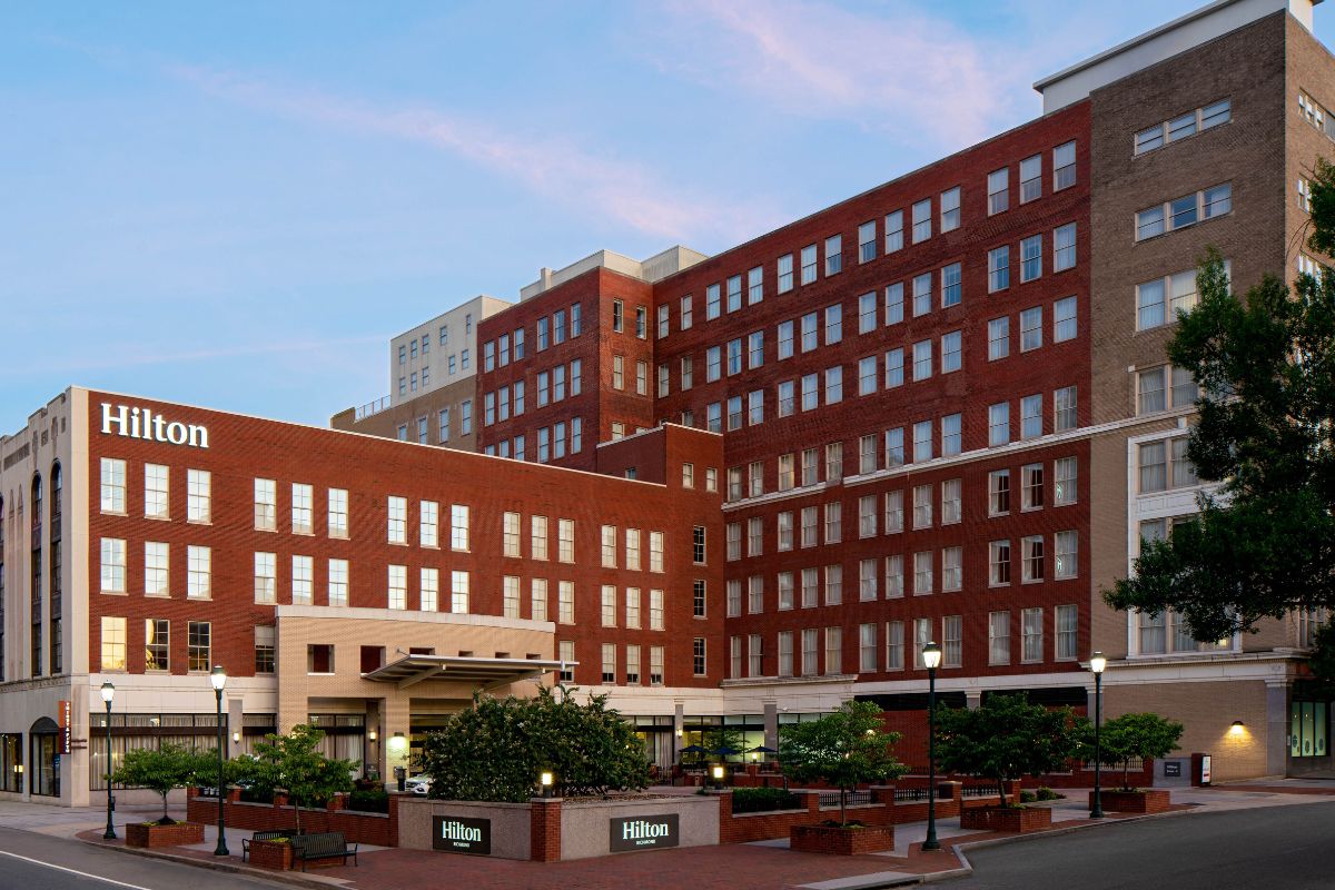 Image of the exterior of Hilton Richmond Downtown (1885), a member of Historic Hotels of America since 2024. Pictured is a large brick building with many windows and greenery.